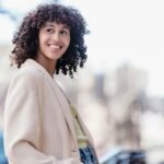Pleasant black female in elegant jacket standing on street and looking away against blurred background