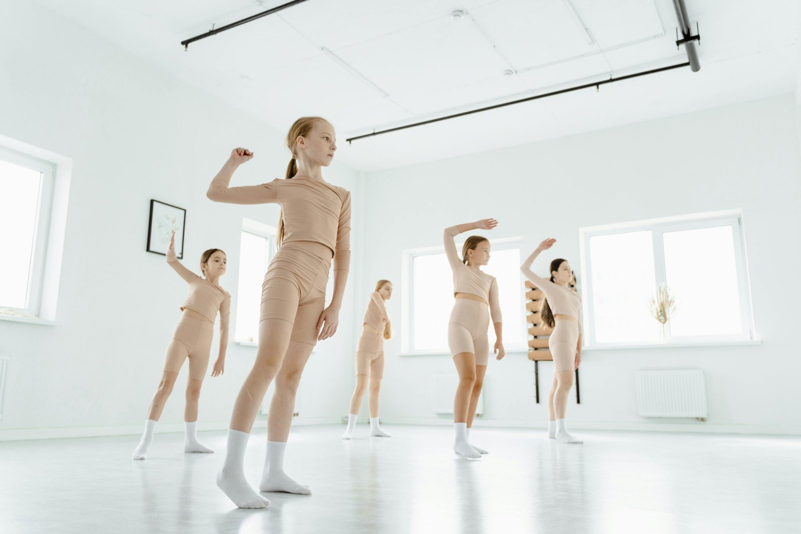 A group of young girls in beige active wear practicing dance indoors in a bright studio.